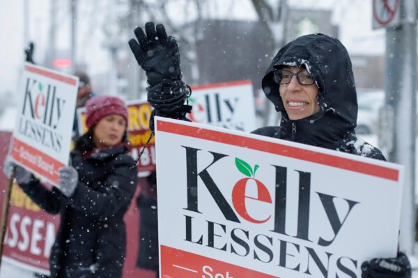 people wave to traffic while holding sings that say "Kelly Lessens" on a snowy day