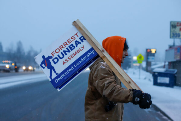 a person walks across an intersection on a snowy day while holding a sign that says "Forrest Dunbar - Assembly - dedicated leadership"