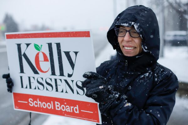 a person holds a sign that reads "Kelly Lessens - school board -" on a snowy day
