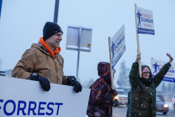 a person smiles while holding a sign that say "Forrest" on a snowy day. Others behind him wave and hold signs that say "Forrest Dunbar - Assembly - dedicated leadership"