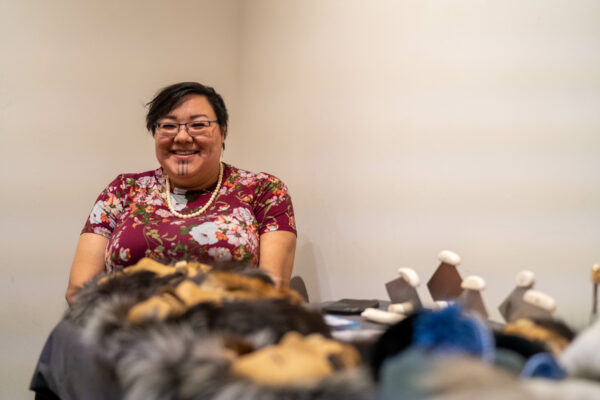 A smiling woman sits next to a table with handmade masks and other crafts.