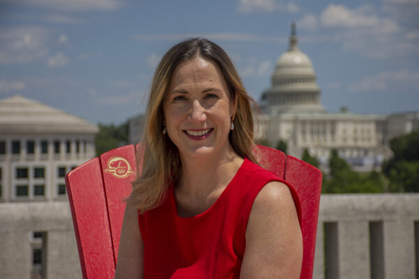 A woman in a red dress sits on a red chair with the slightly unfocused background of the U.S. Capitol building