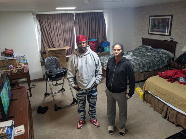 Two african american adults stand in a hotel room next to two queen beds and a high chair