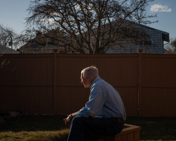 An elderly man sitting on a wooden planter in a back yard