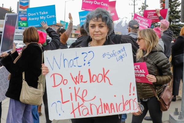a person stands with a sign that reads "what? do we look like handmaidens?" in front of people at a rally