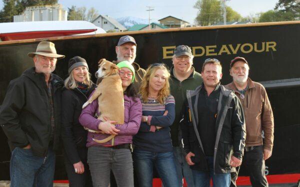 A group photo of eight people and a dog posing on a dock in front of the Endeavor