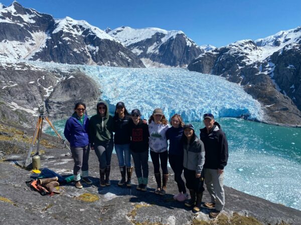 A photo of a group standing at the edge of a hill with a glacier's terminus in the background