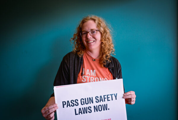 A woman with an orange shirt on holding a sign that says pass gun safety now.