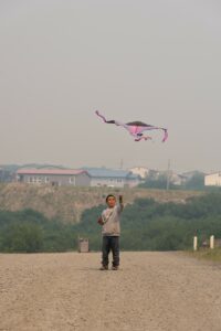 A boy flies a pink kite on a dirt road with dense smoke in the air and a village in the background
