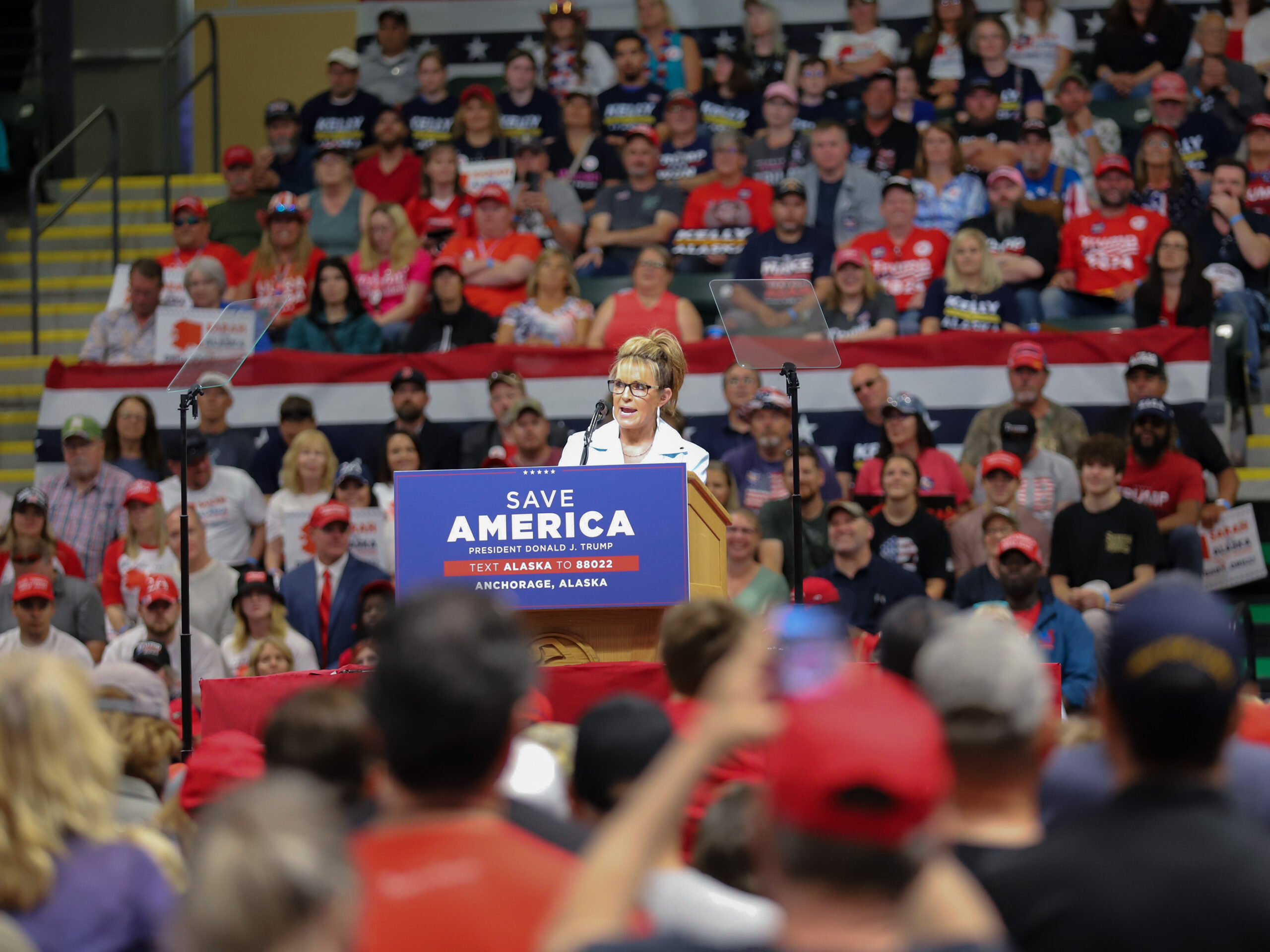 Sarah Palin speaks at a podium, a crowd surrounds her