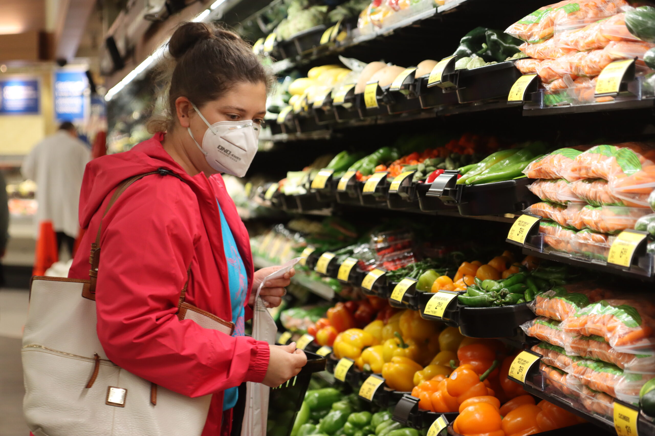 A woman in a pink jacket holds a plastic bag in front of produce shelves at a grocery store.