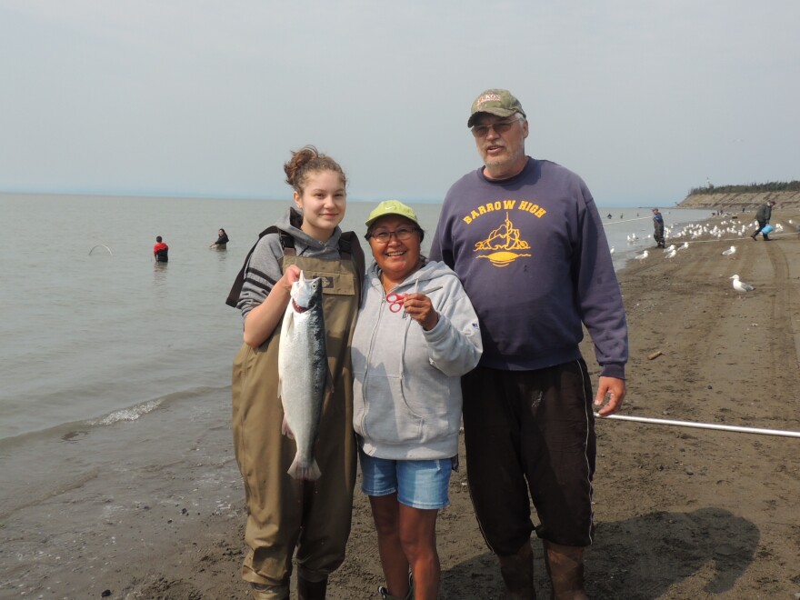 three people post for a photo on a beach, one of them in waders is holding a salmon