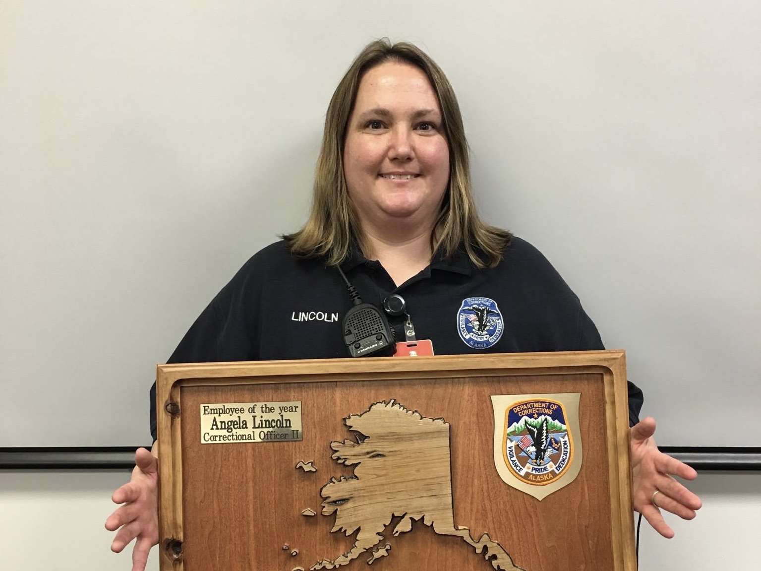 A woman smiles, facing the camera and holding a wooden award that says "employee of the year, Angela Lincoln, correctional officer II" and has the shape of Alaska on it.