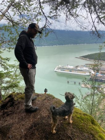 a man and his dog stand on a mountainside, overlooking a docked cruise ship