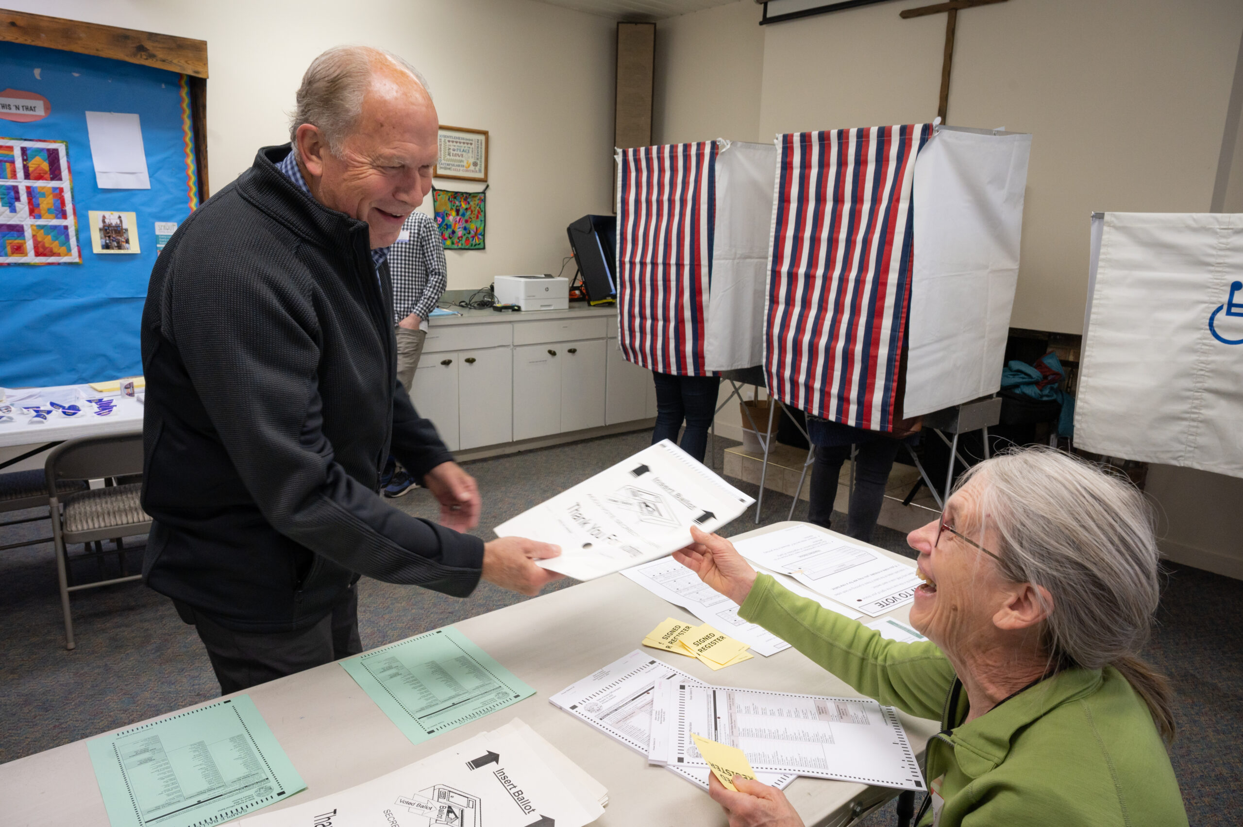 A man in a black coat receives a voting ballot from a woman in a green shirt.