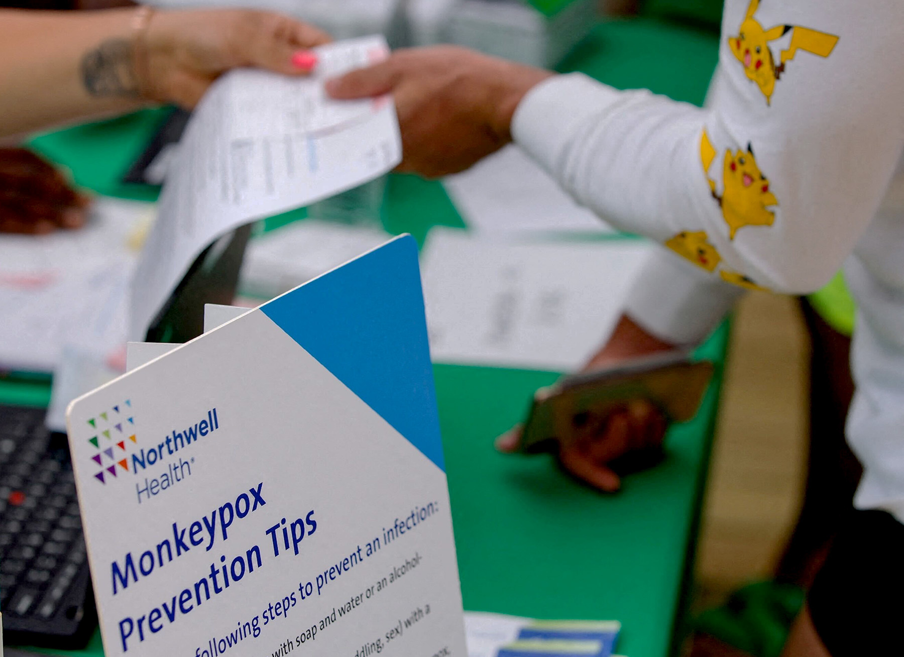 a piece of paper exchanges hands in back of a sign that say "monkeypox prevention tips"