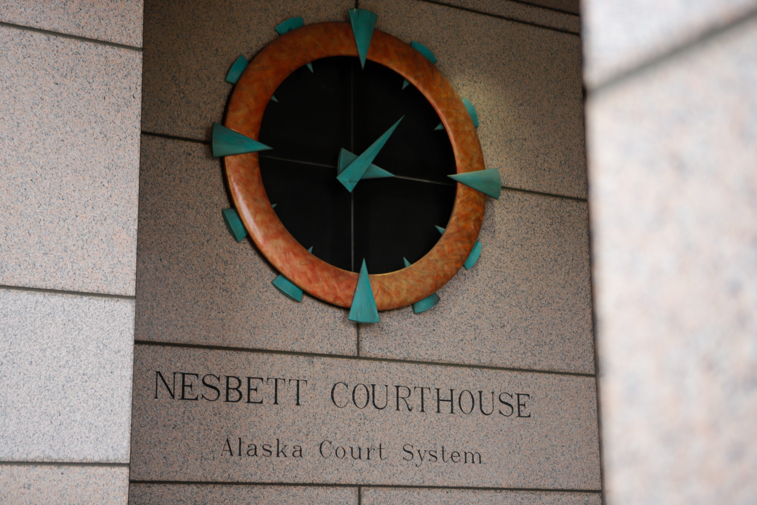 An analog clock with a black center surrounded by orange stone and with turquoise hands and hour markers reads 3:07.