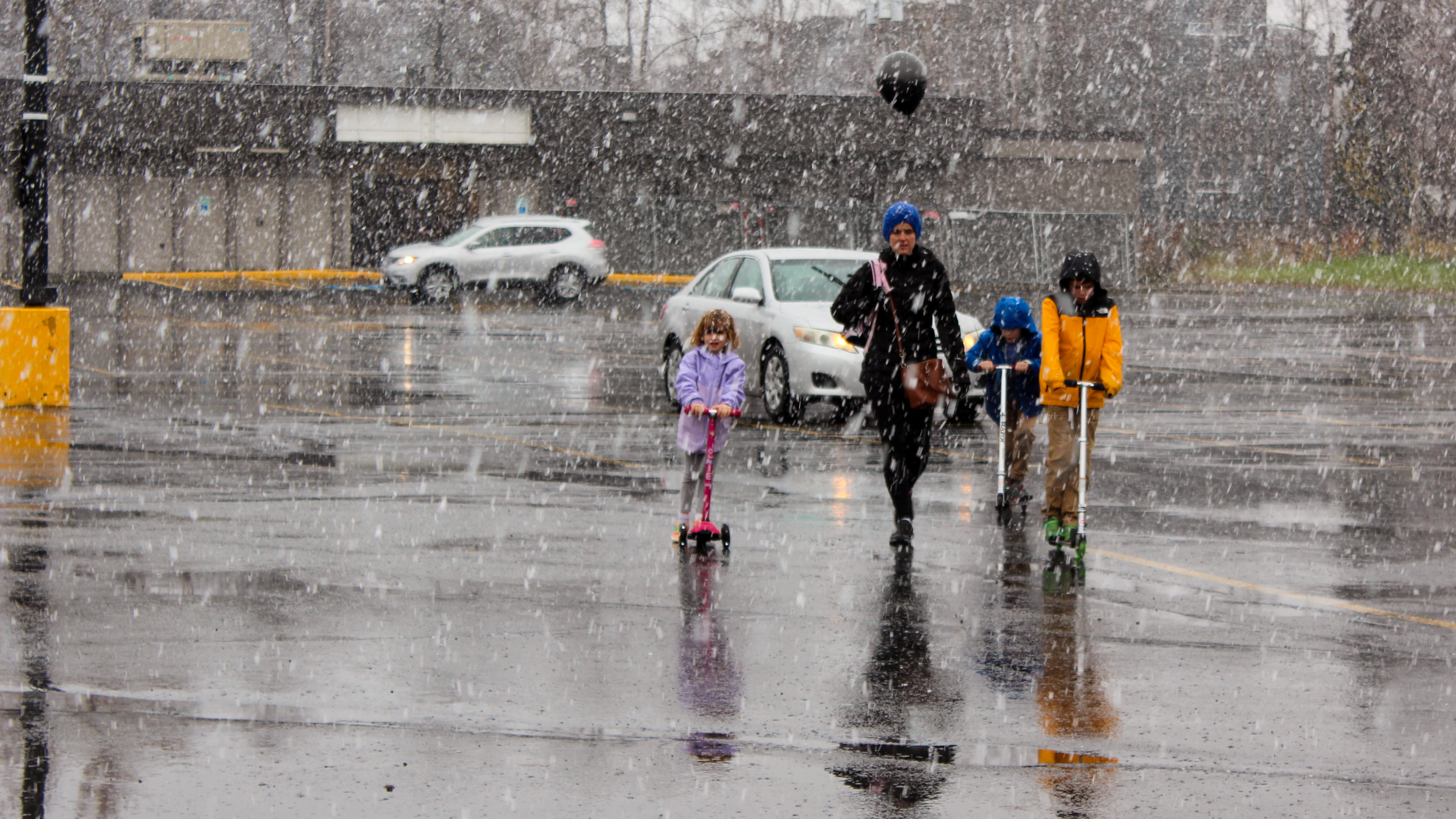 A mother holding a ballon walks through a parking lot while three children in jackets ride scooters.