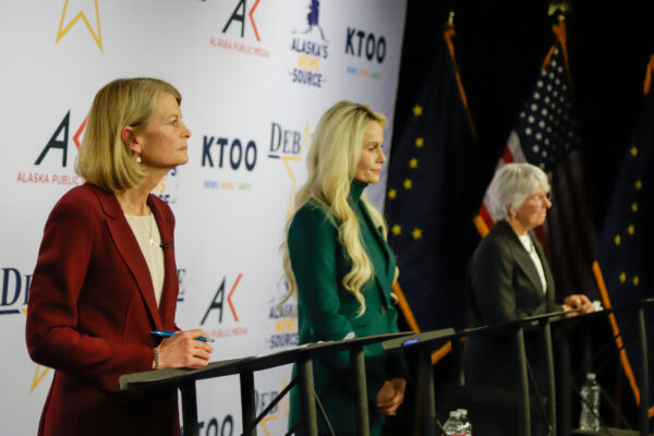 Closest to the camera is a woman in a red blazer, in the middle is a woman in a green suit and furthest from the camera a woman in a black suit. All three stand behind podiums while listening to a question during a political debate.