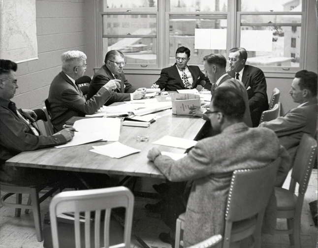 A black and white photo of eight men in suits sitting around a table with papers.