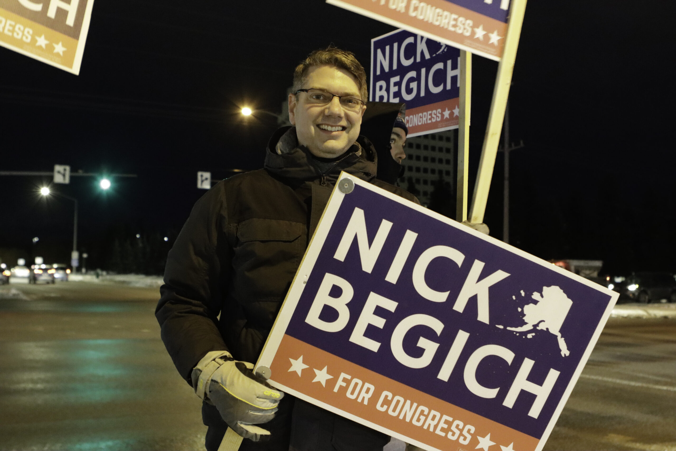 a man stands outside holding a sign that says Nick Begich for Congress