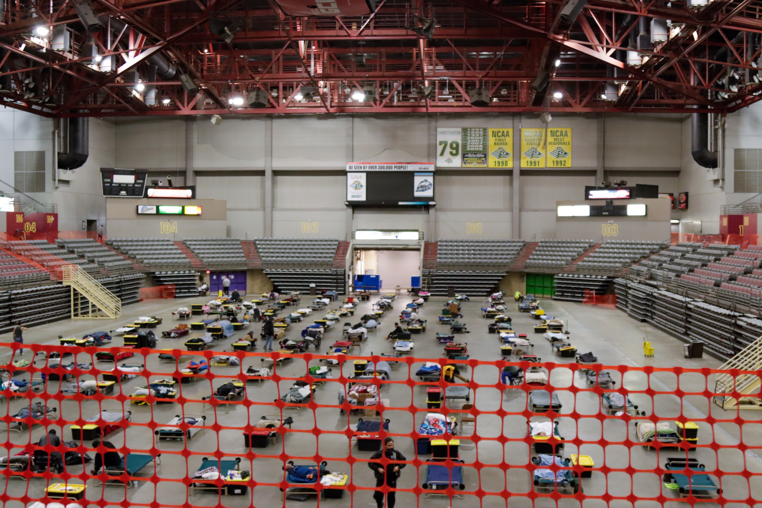 Rows of cots are organized on the floor of an arena.