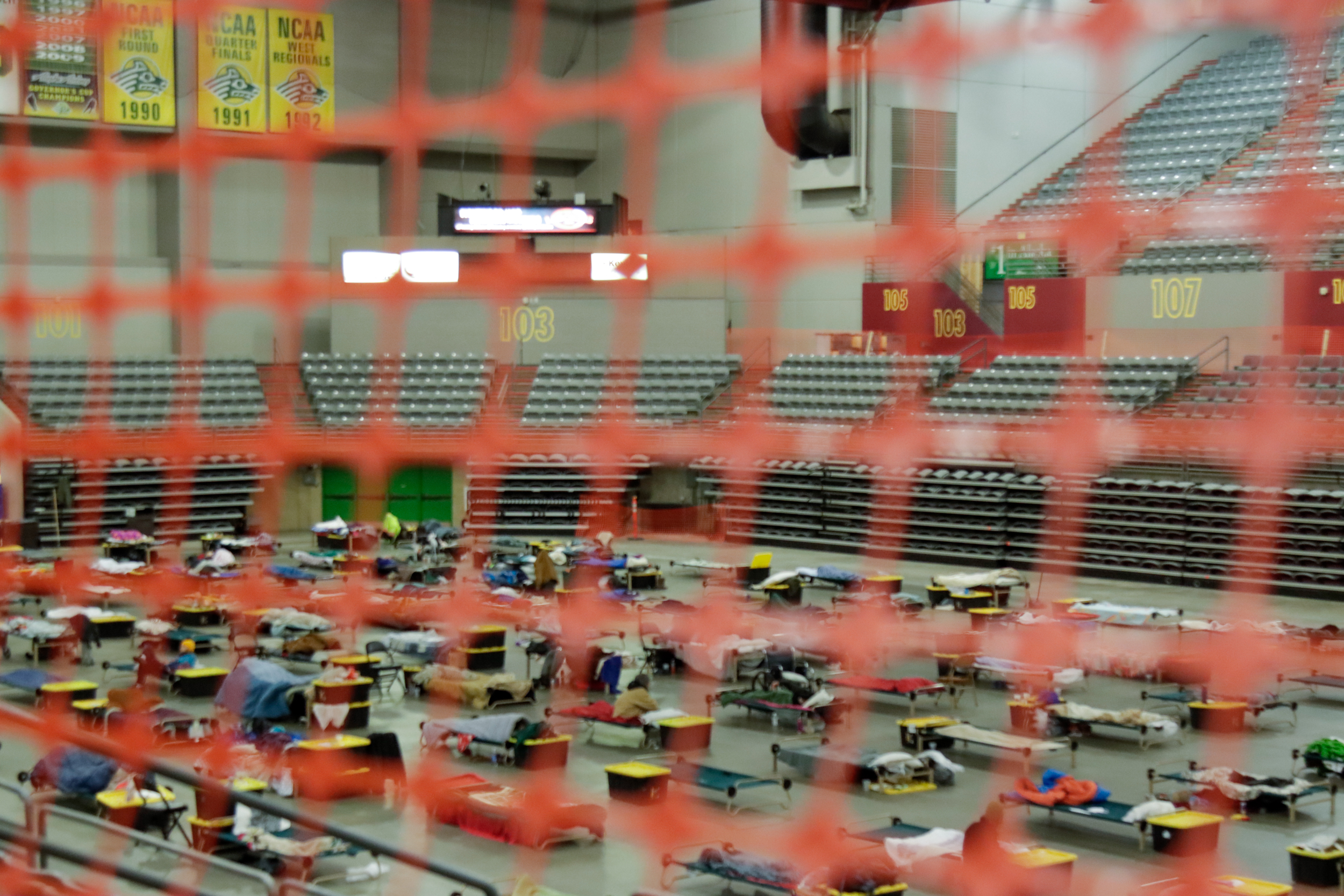 Cots and totes are organized on the floor of an arena.