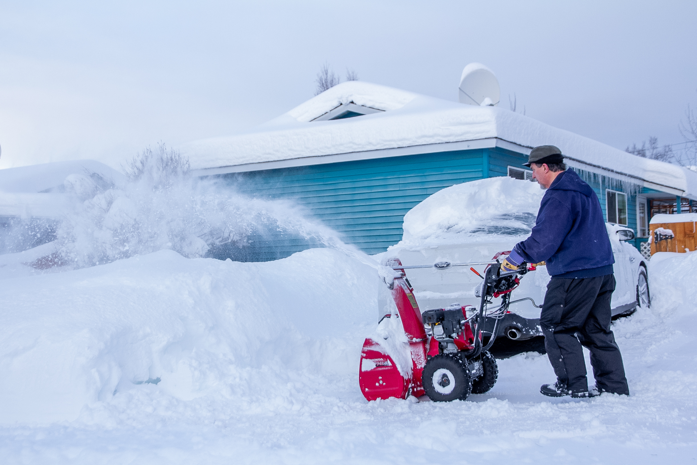 A man in a hat pushes a snow blower spraying snow.