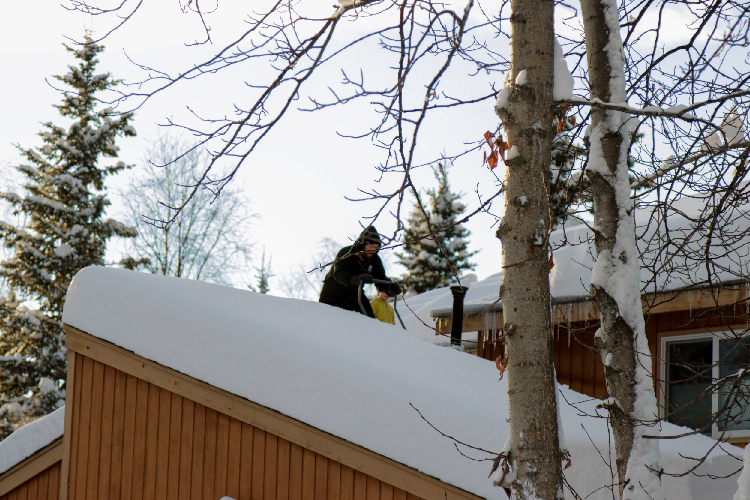 A man uses a push shovel on the snow covered roof of a house.