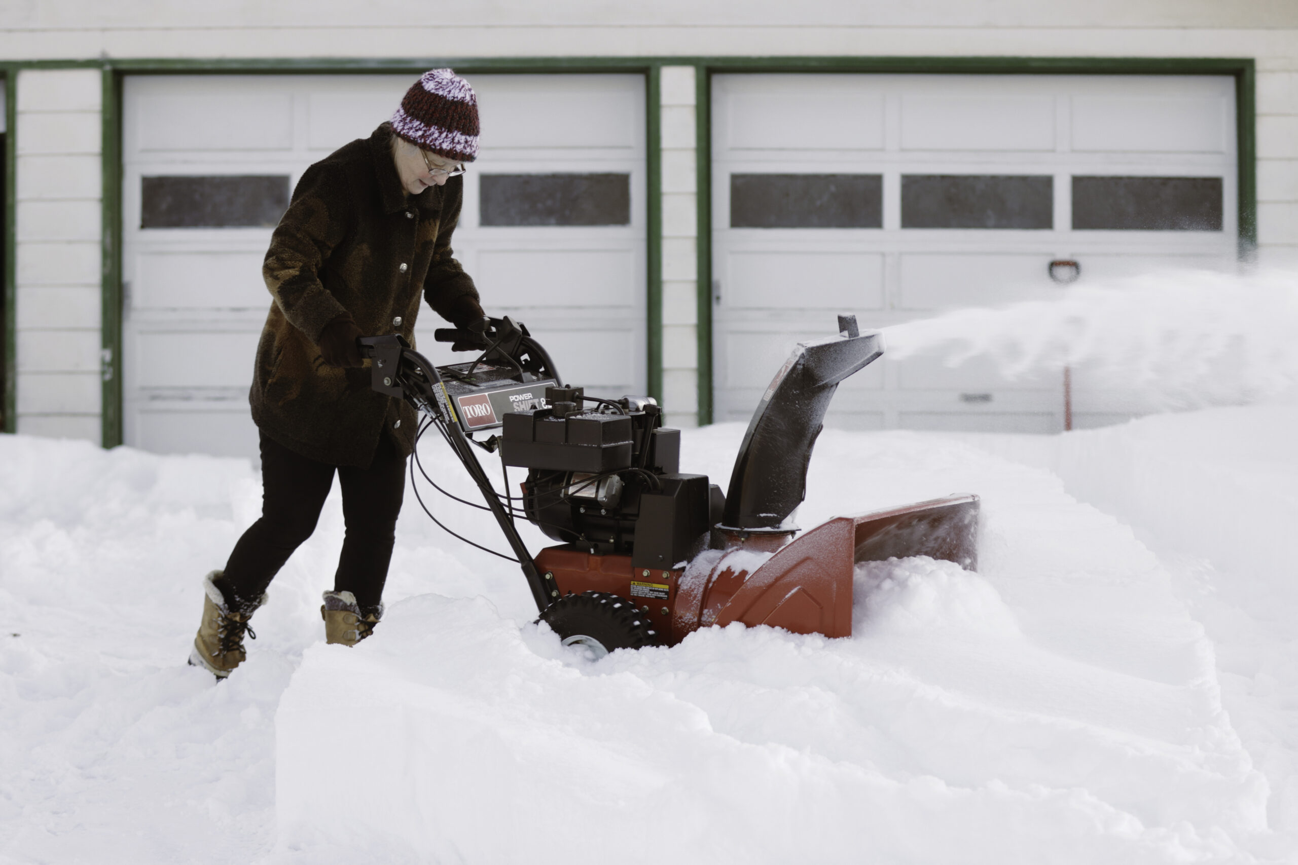 a woman pushes a snowblower in a snowy driveway