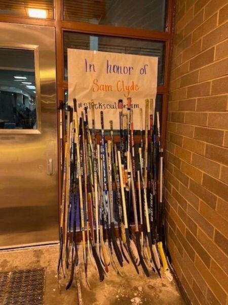 Hockey sticks lined up outside, in front of a sign that says "In honor of Sam Cylde)