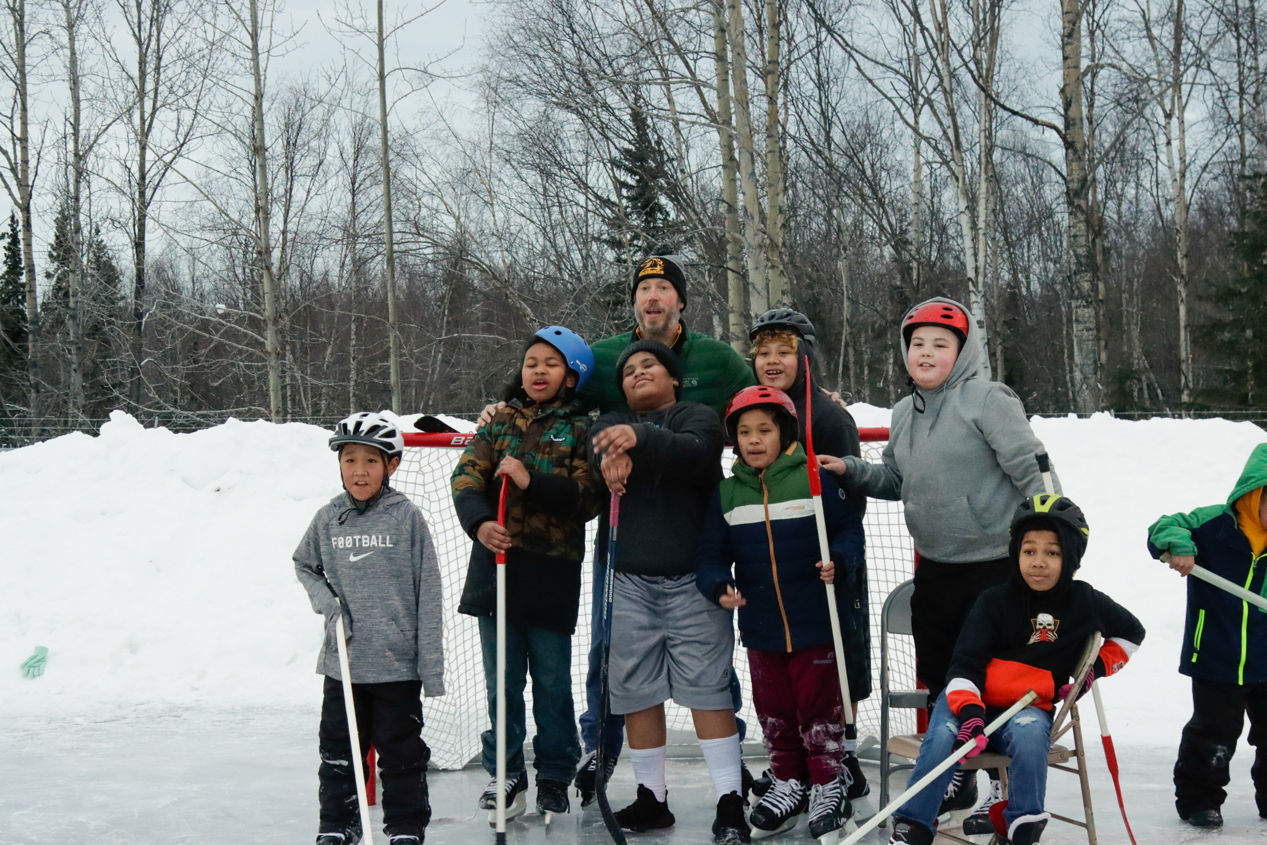 A group of children pose with their teacher in front of a hockey goal on an ice rink.