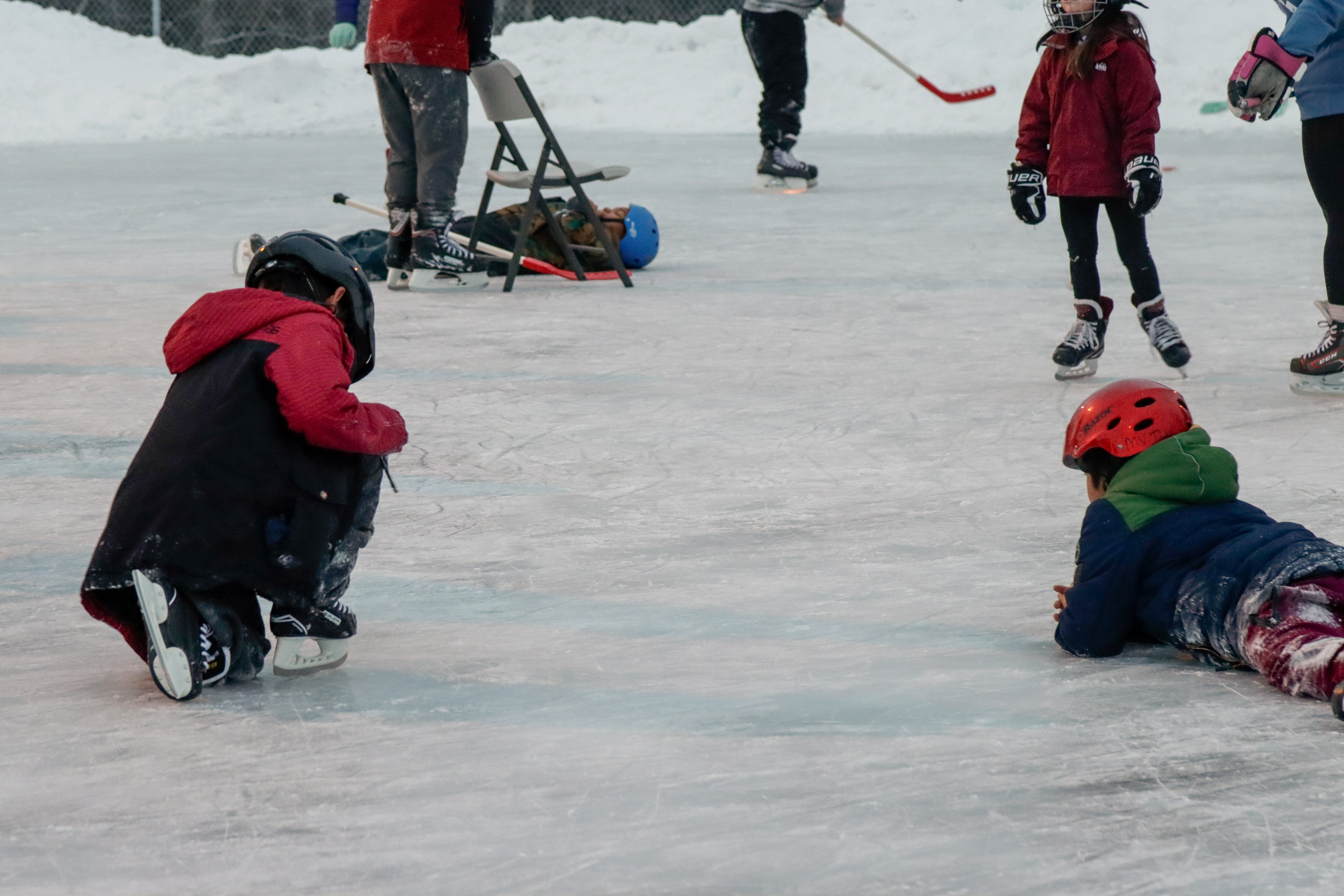 Two children in helmets and skate sit on an ice rink while others skate around them.
