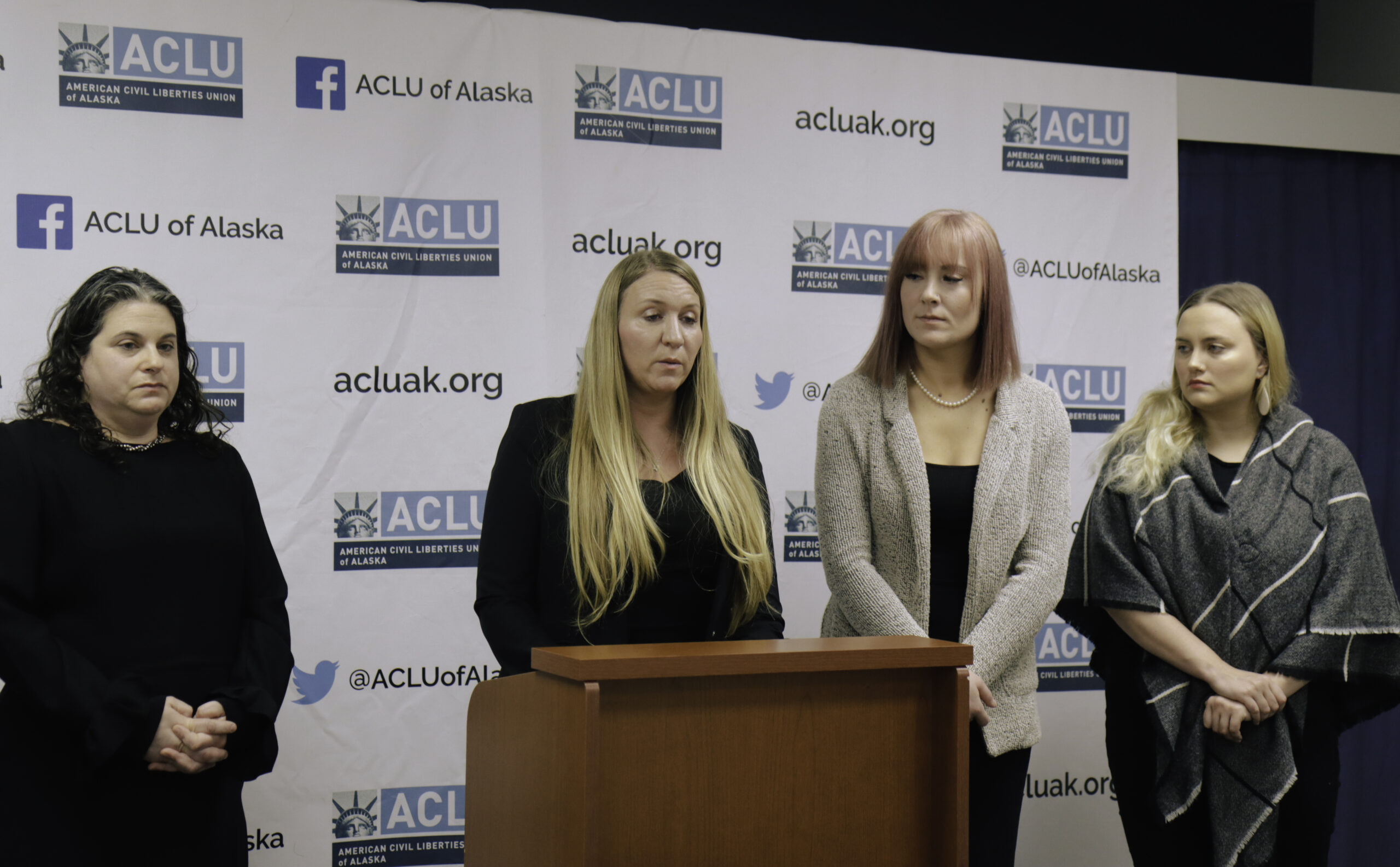 Four women stand behind a podium discussing ACLU of Alaska Prison Project. A woman with long blonde hair talks in front of press conference.