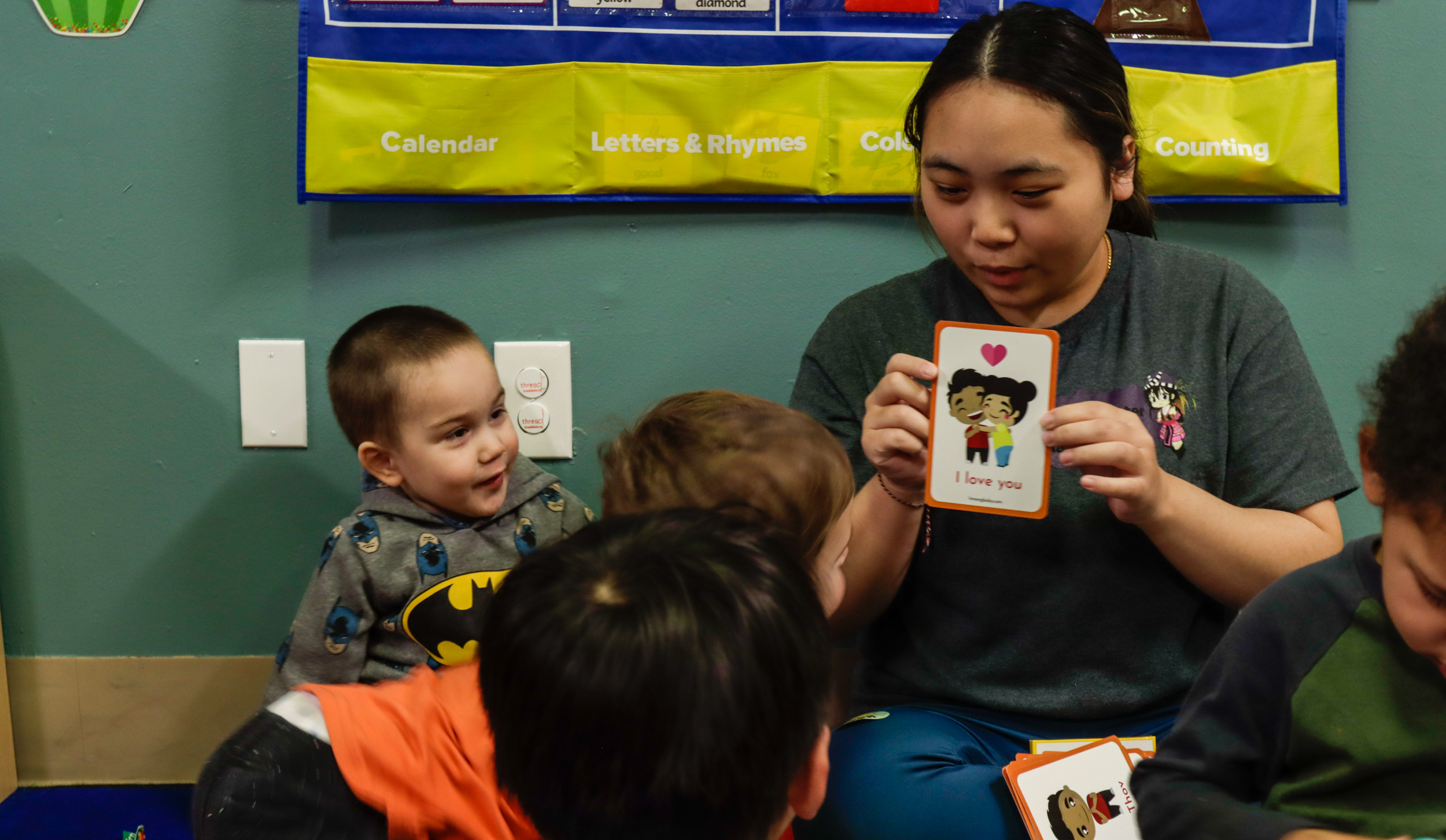 A woman shows a flash card of two figures hugging to a room of toddlers.