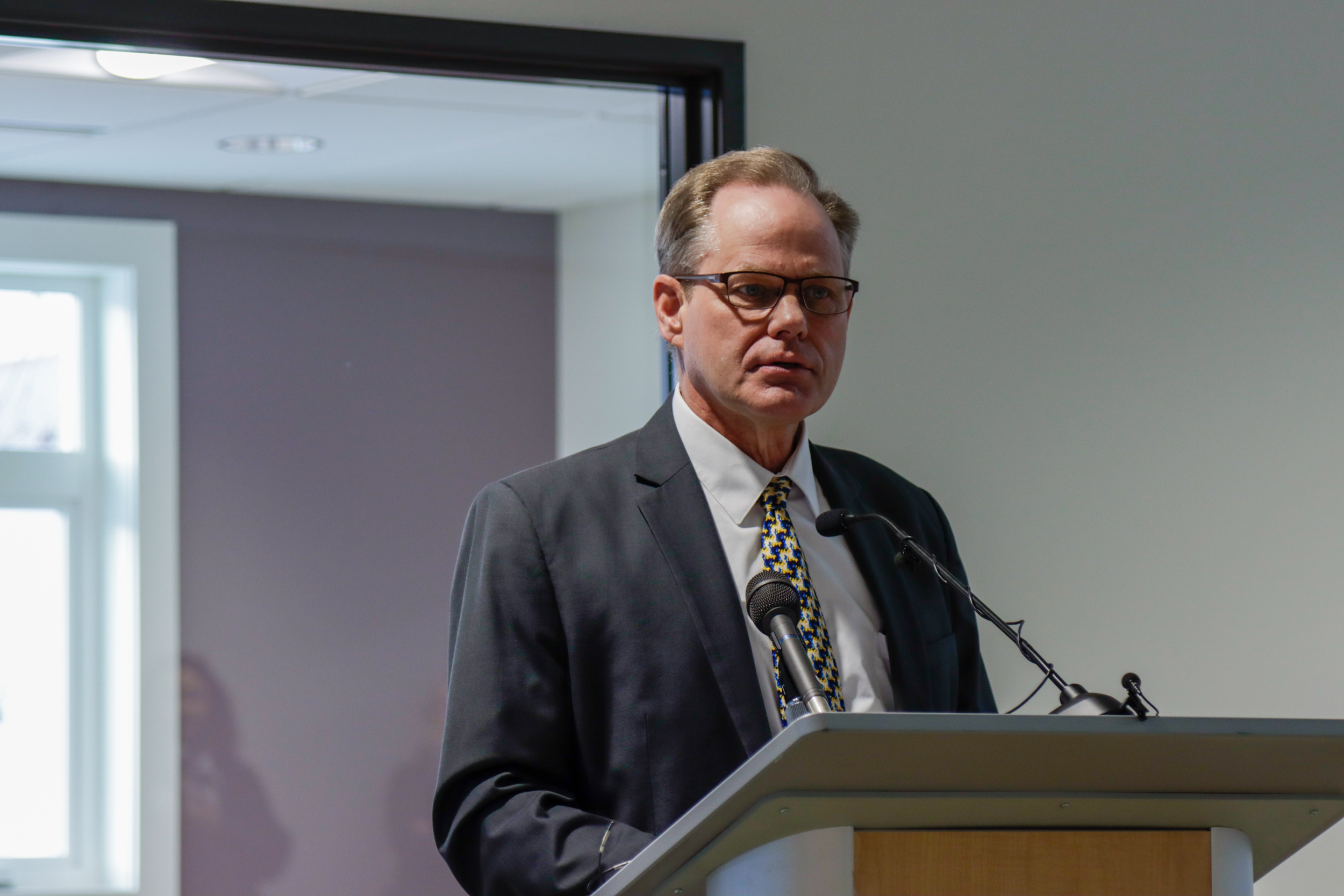 A man in a suit and glasses speaks from behind a podium.