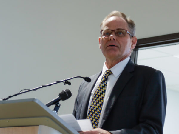 A man in a suit and glasses speaks behind a podium.
