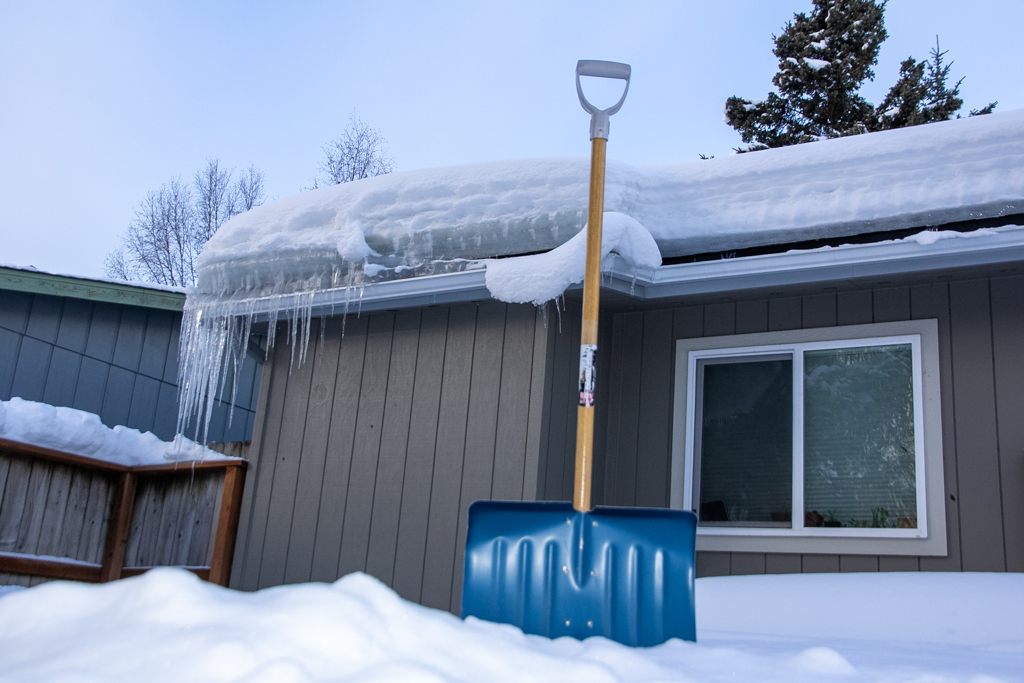 A snow shovel sits in front of a house with a snowey and icey roof.