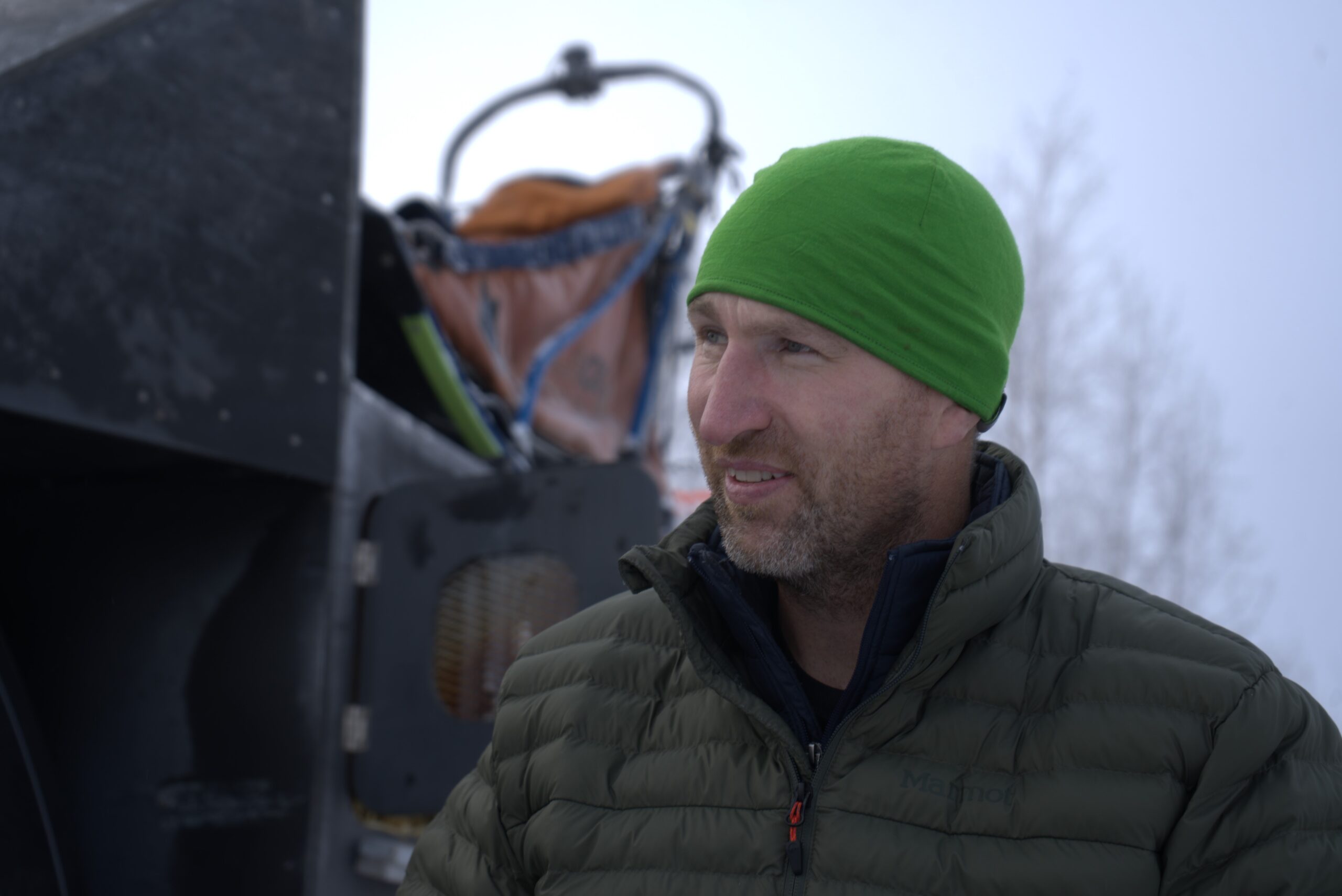 A man with a green hat and down jacket in front of a gog sled strapped to teh roof of a truck