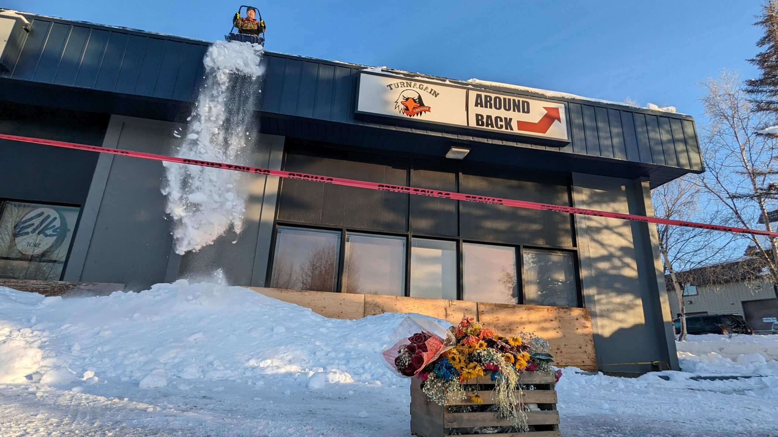 a man pushes snow off the roof of a gym with a box of flowers in front