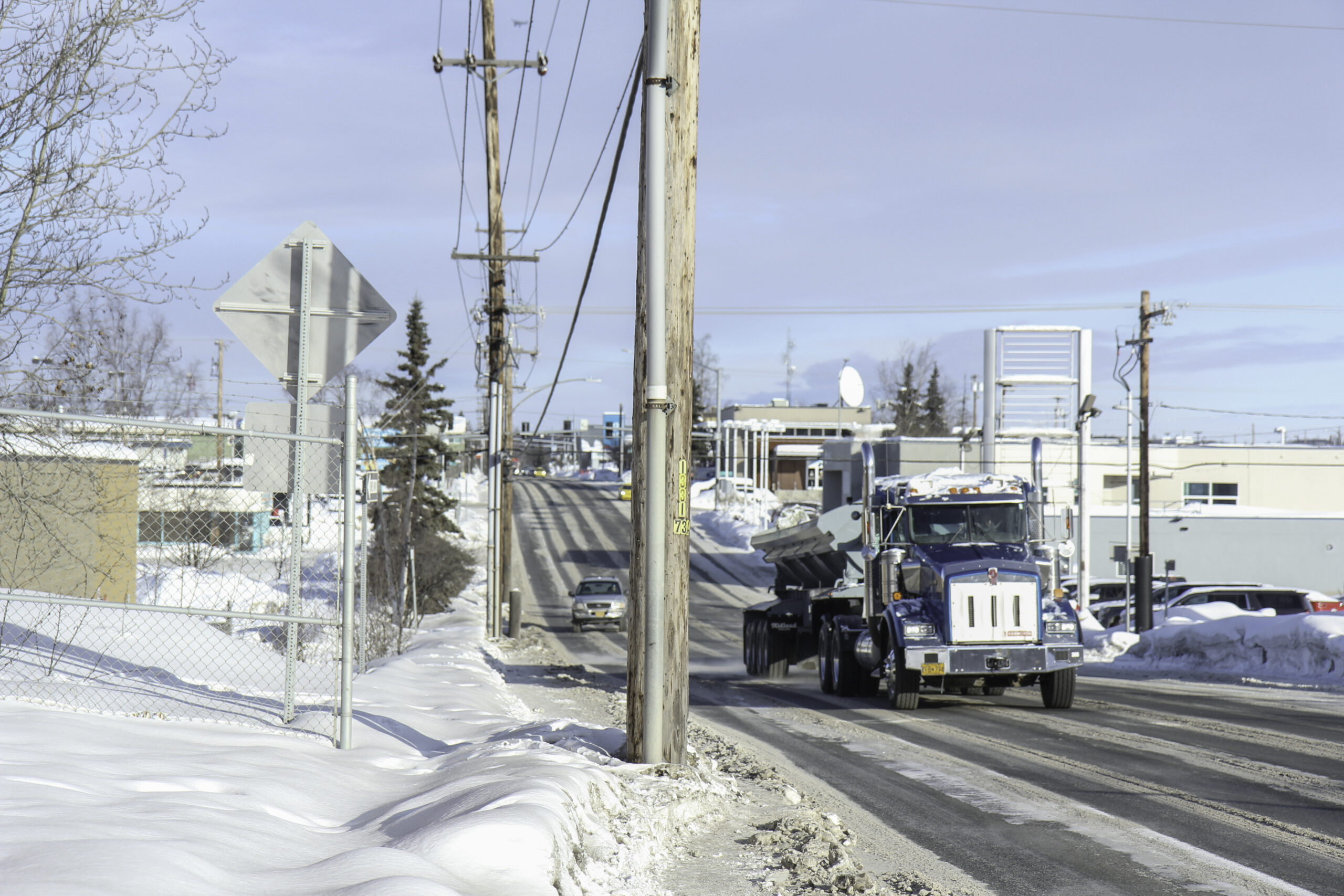 A sense of place photo in the winter where a semi truck is on the right side of the photo where the road is and utility poles obstructing pedestrian sidewalks on a snow filled street.