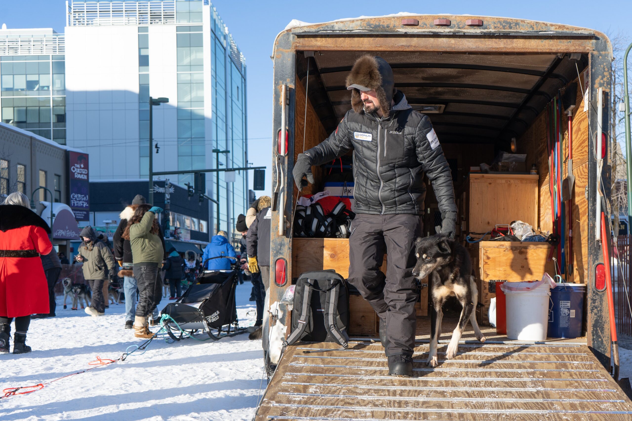A man in black winter gear pulls his dog down a ramp toward a dog sled