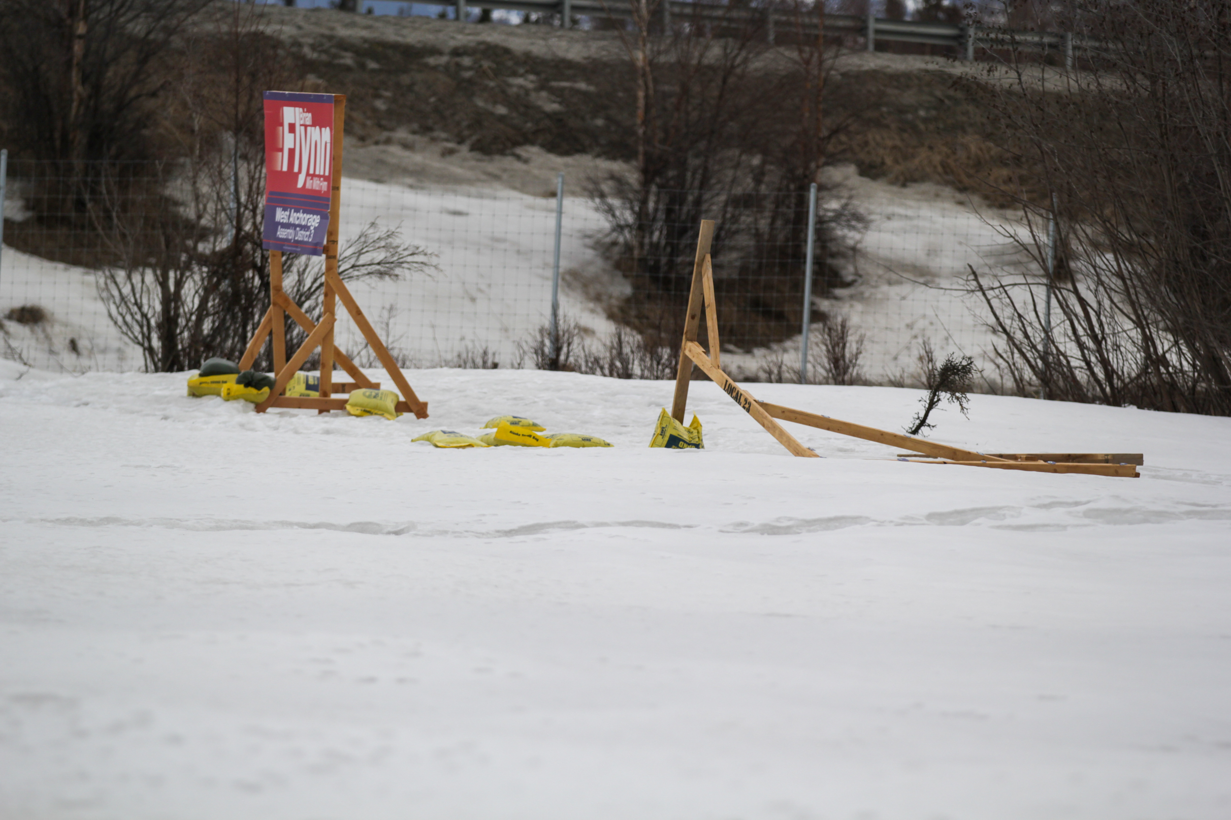 wooden sign holders and sand bags are strewn on the snow-covered ground. In the background, a wooden frame holds another political sign.