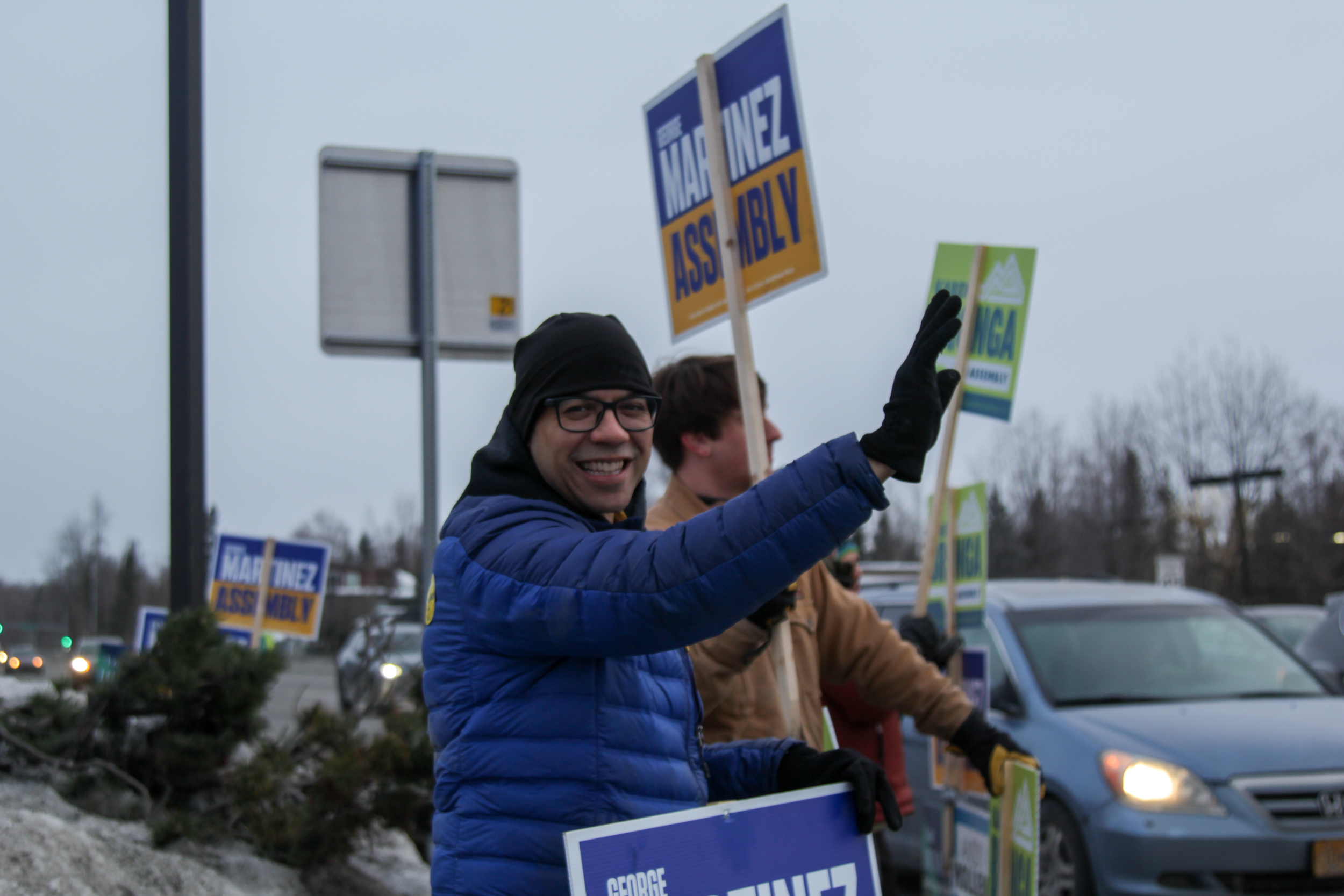 A man in a blue jacket waves to traffic while holding a political sign.