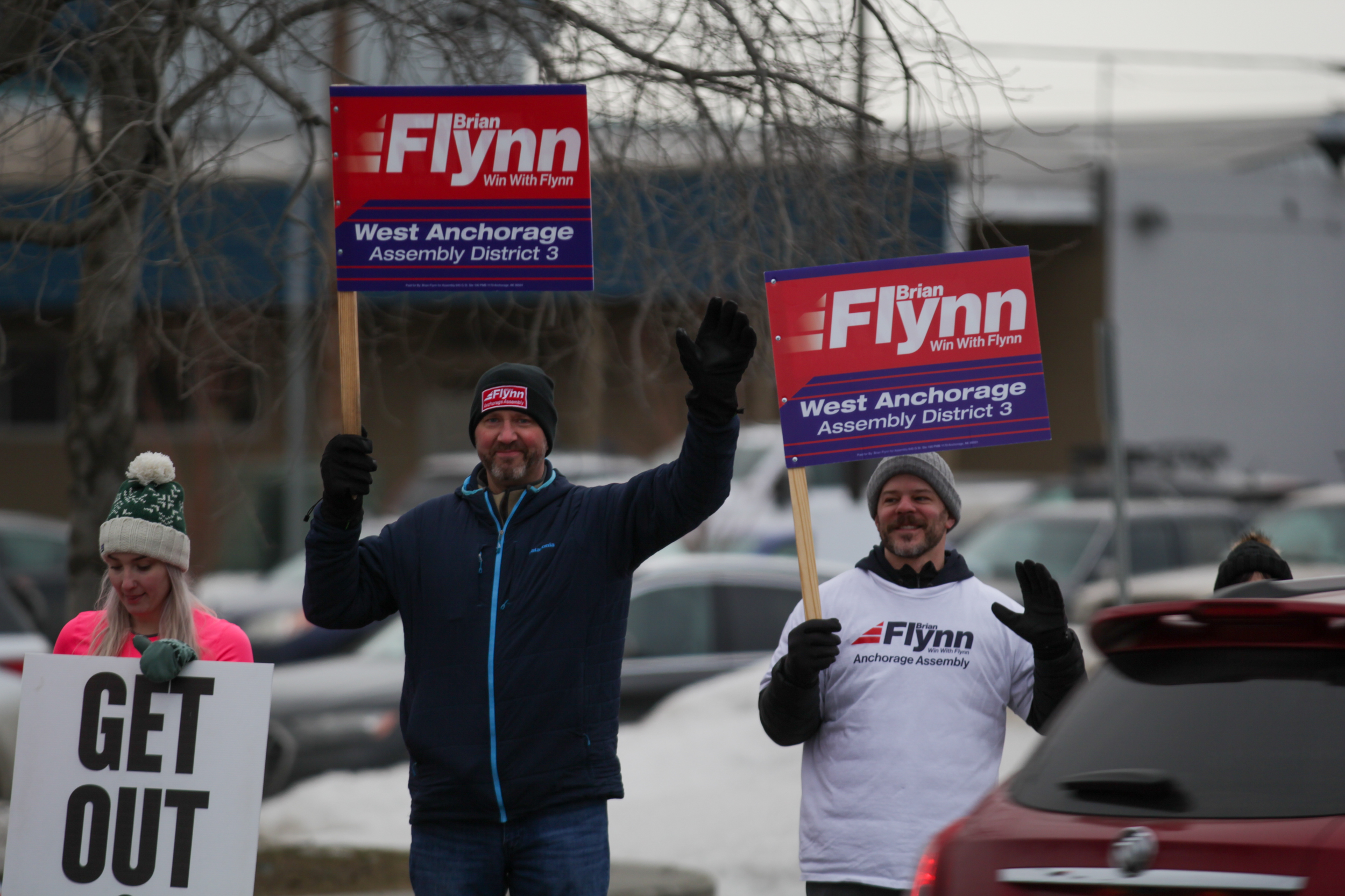 Two men holding political signs wave to traffic at an intersection.