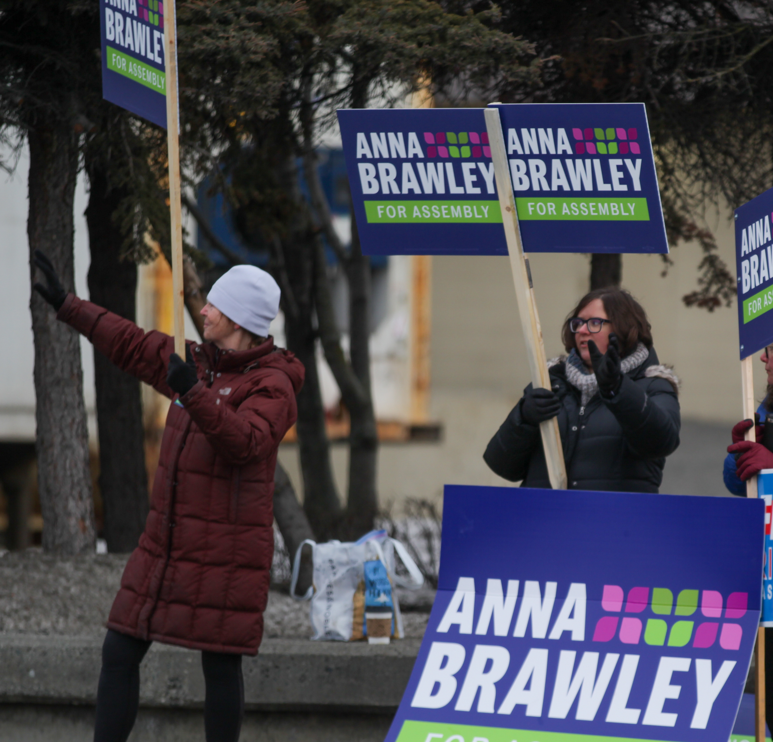 Women in winter coats wave political campaign signs at an intersection.