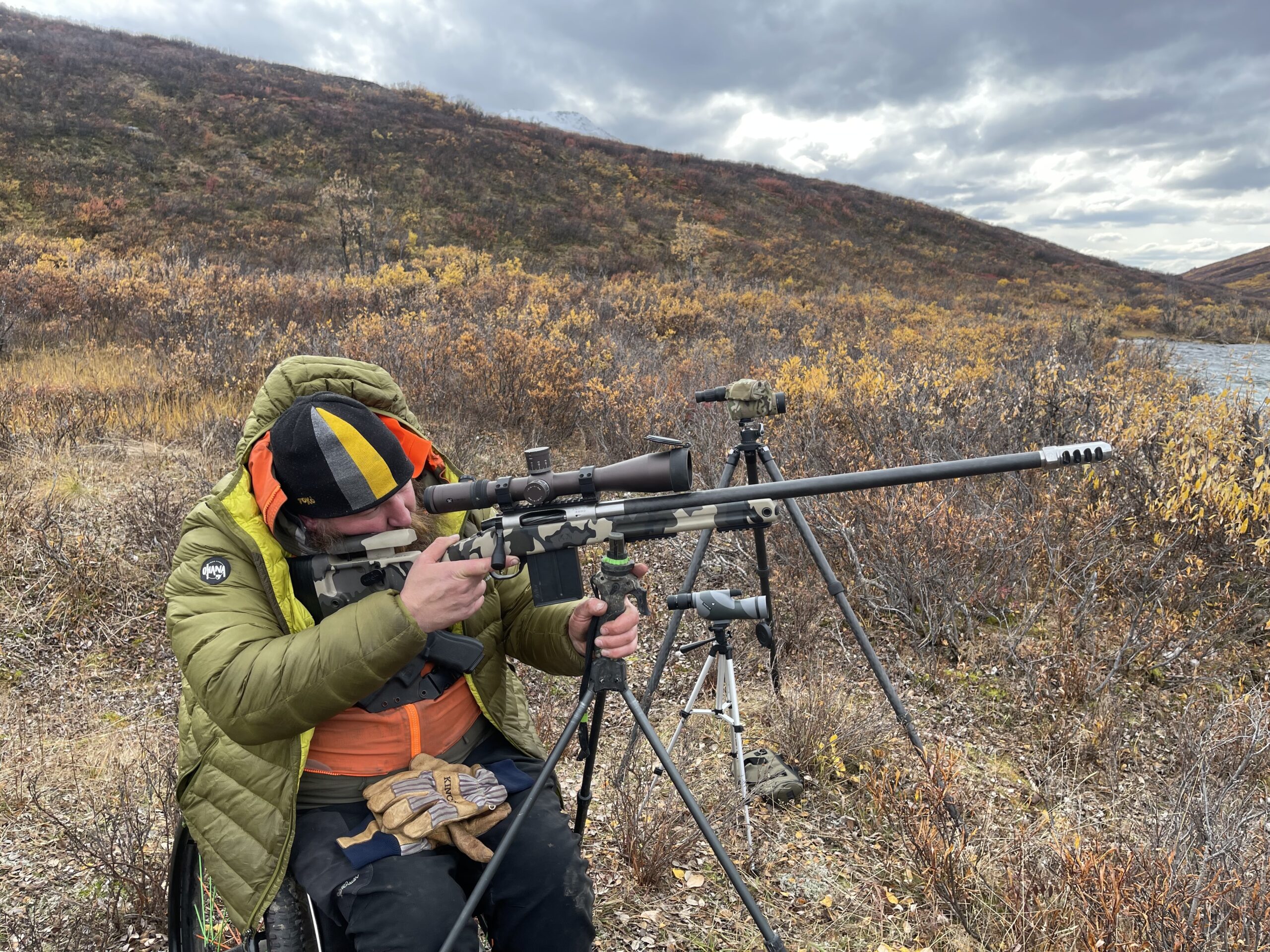 A seated man in a green puffy coat looks through the scope on a rifle, which is mounted on a tripod, with tundra shrubs in the background.