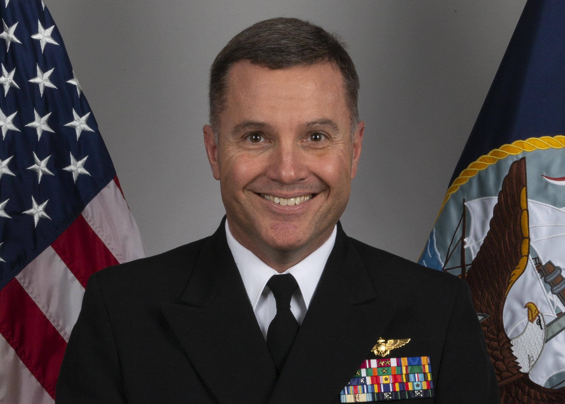 A white man in a dark blue uniform looks directly into the camera for an official portrait with the U.S. flag and U.S. Navy flag on each side.