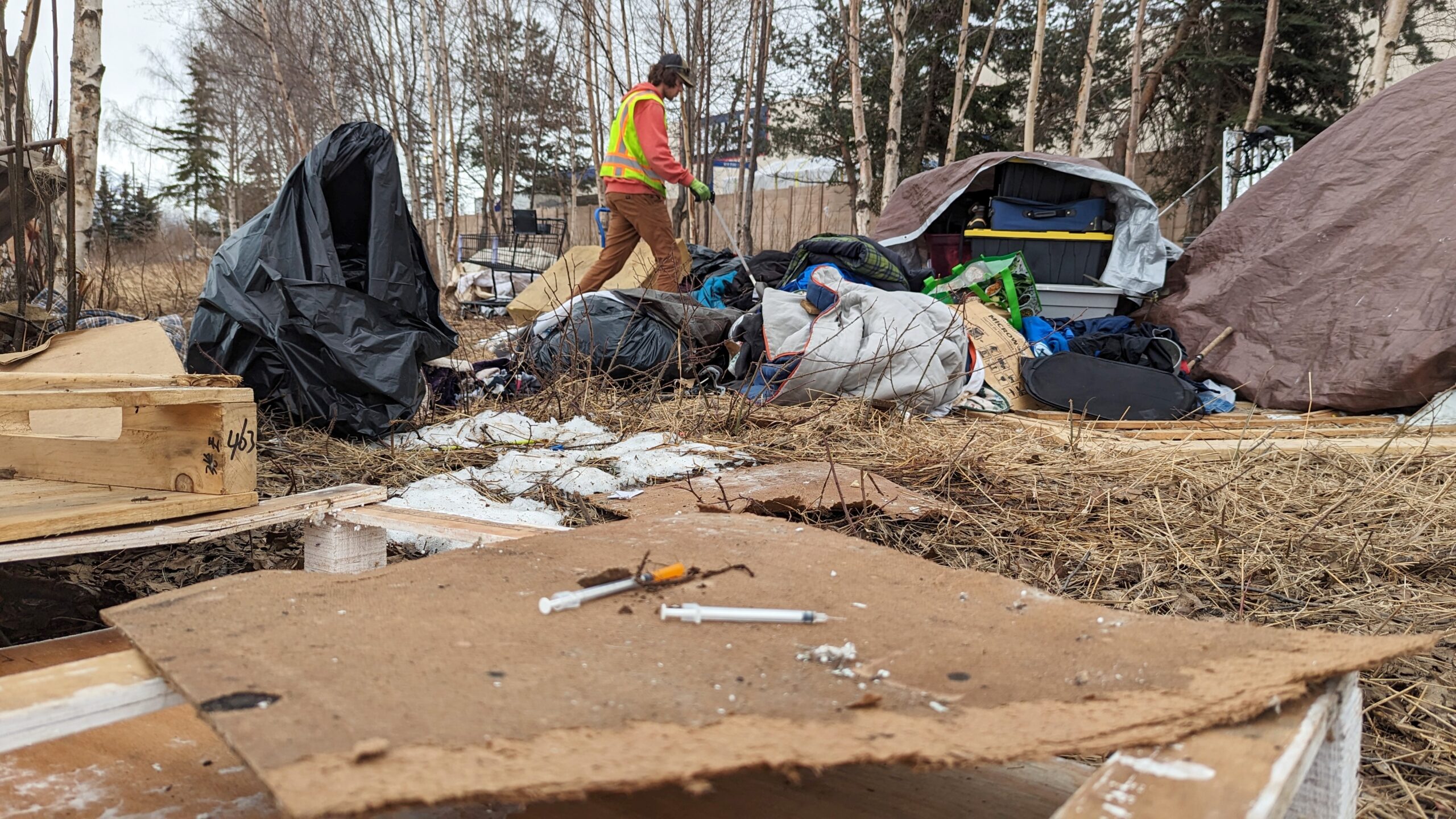 A man in a reflective vest picks uses a trash picker around tarps, tents and needles.