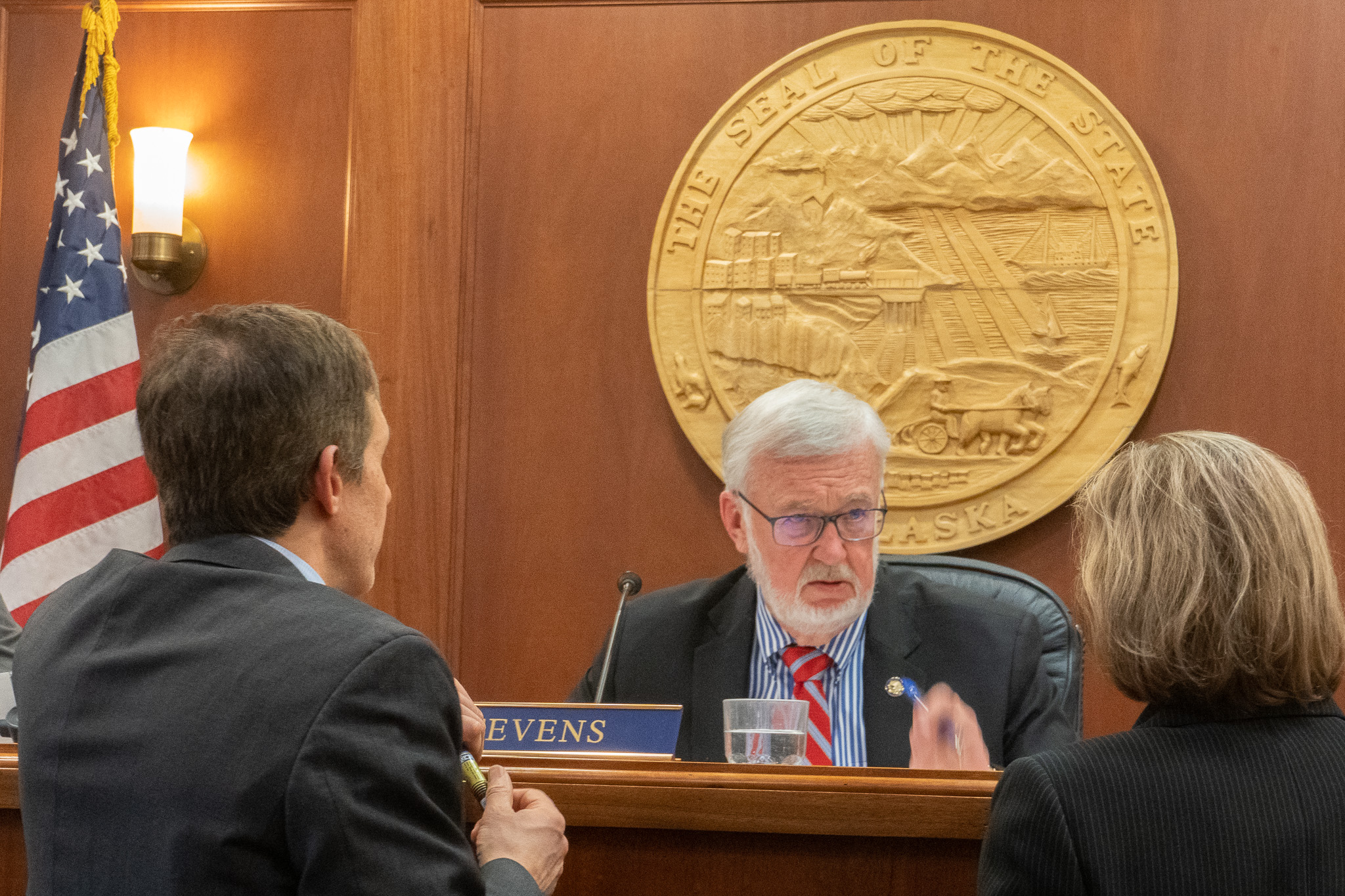 A white bearded man sits at a desk in front of a gold seal while two people talk to him.